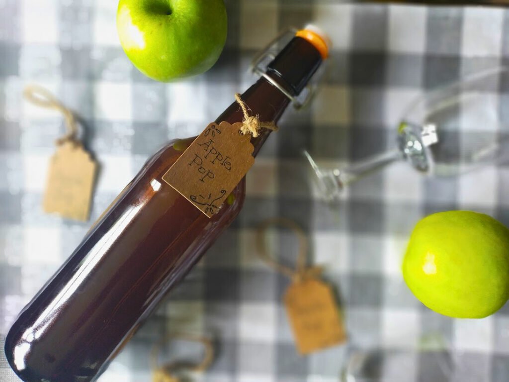 Amber bottle of apple pop lying on its side on a grey and white check cloth surrounded by a wine glass, brown tags and two green apples