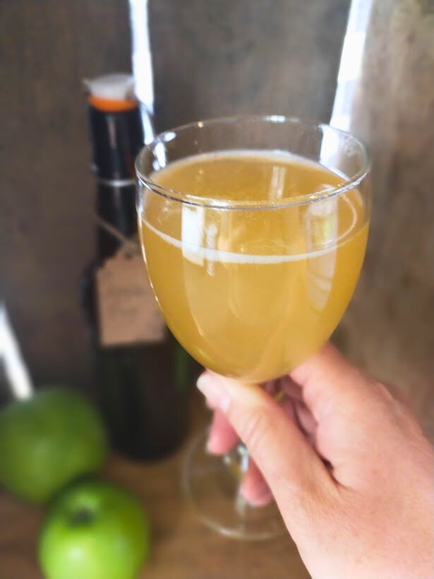 Hand holding a glass with apple pop drink in. Amber bottle and two green apples sitting in a wooden box in the background.
