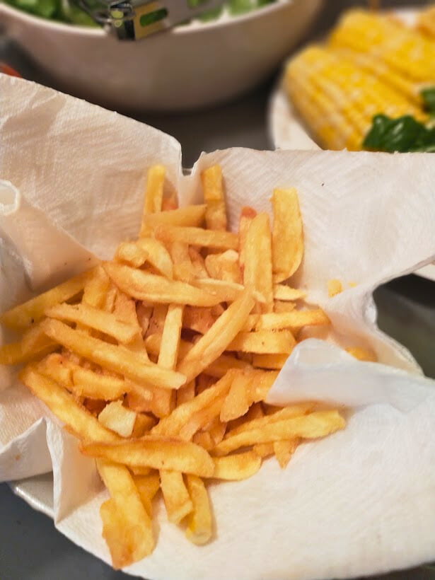 Homemade fries on a paper towel lined plate, Bowl in background and corn on the cob.