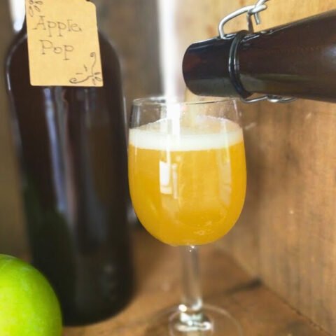 Pouring apple pop into a glass, bottle and apple in the background, wooden backdrop