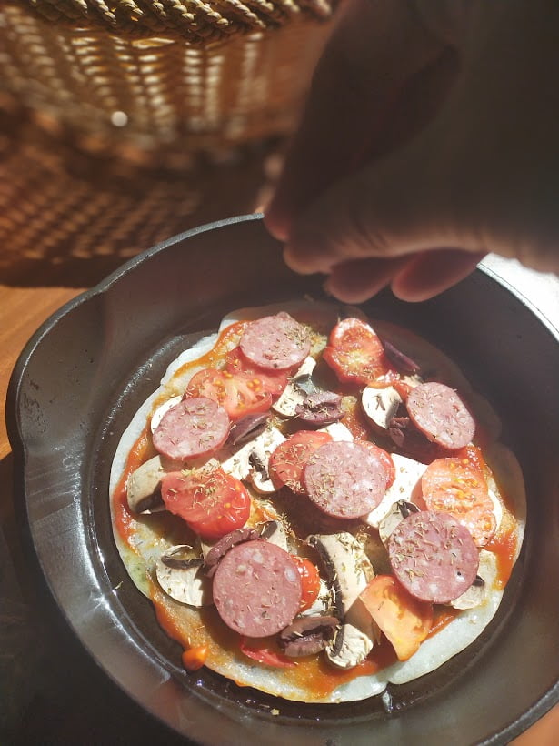 hand sprinkling dried herbs to a pizza in a cast-iron skillet