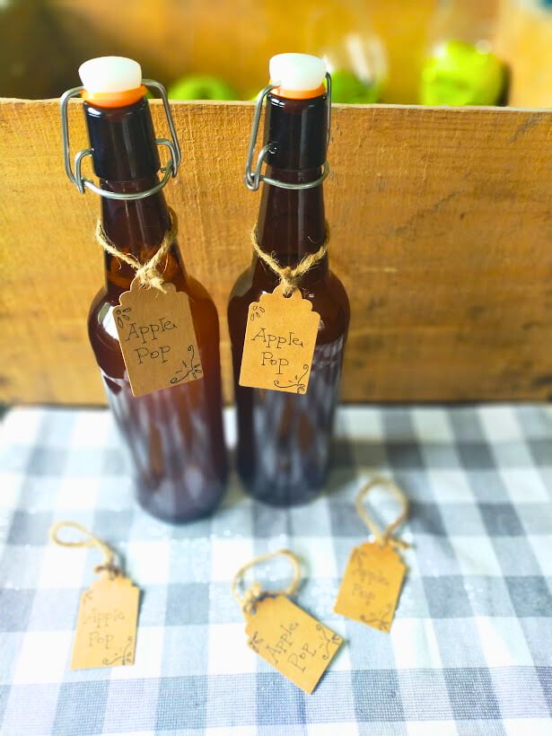 Two amber bottles sitting on a grey and white checked cloth. Wooden box with green apples in the background