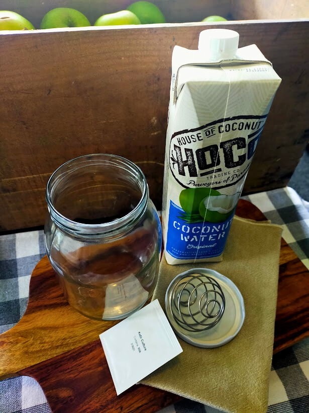 Glass jar, mixing ball, coconut water and kefir culture sitting on a wooden board