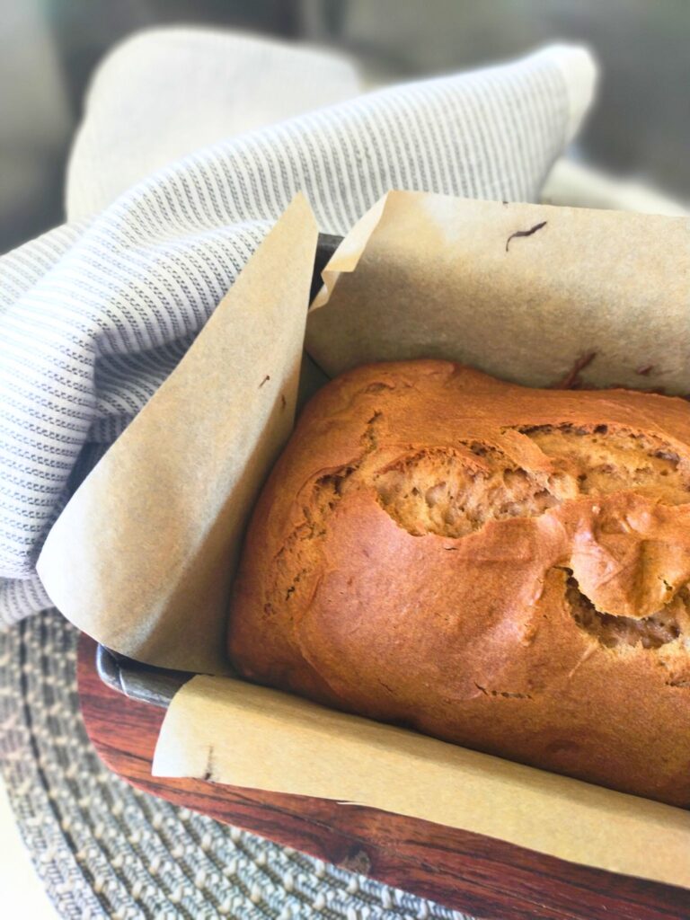 Baked gluten free gingerbread loaf in a loaf tin with baking paper sitting on a wooden board with a blue and white tea towel in the background.