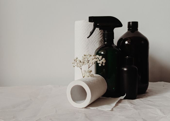 Amber spray bottles in a variety of sizes sitting on a white background with paper towels, toilet paper and white flowers.