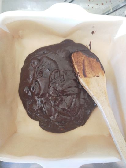gluten free brownie batter in the lined baking dish with a wooden spoon spreading it out.