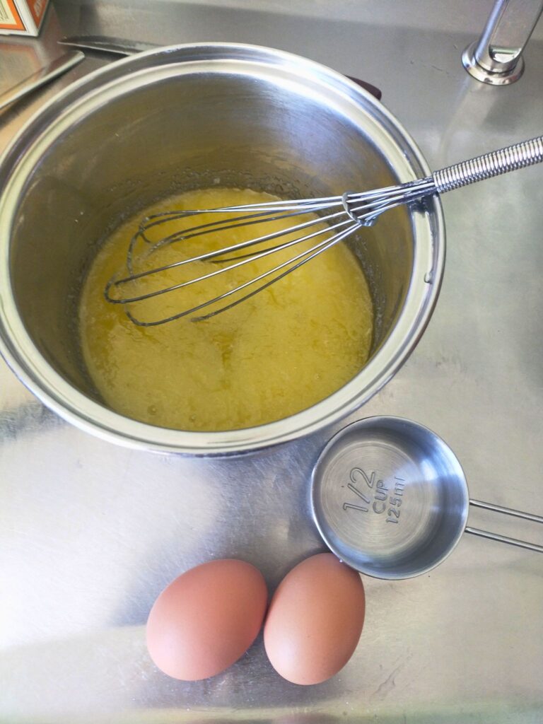 Brownie ingredients in a pot with a whisk, two eggs and a measuring cup in the foreground.