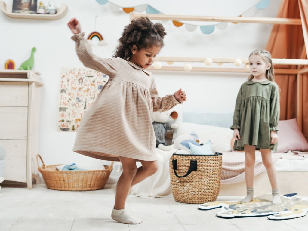 Two young girls in dressing dancing in the bedroom surrounded by toys and bedroom furniture.