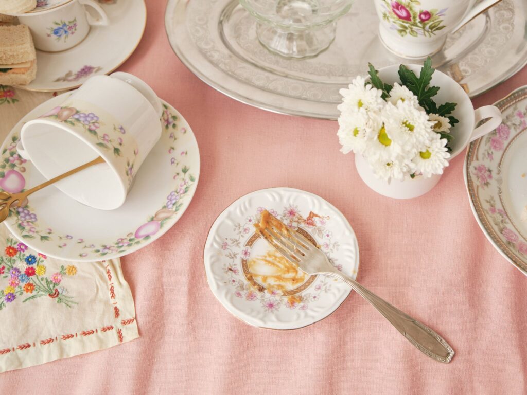 A table set with old cups, saucers and plates on a pink tablecloth. One of the cups has daisy flowers in it. There's a plate with a fork and some leftover food.