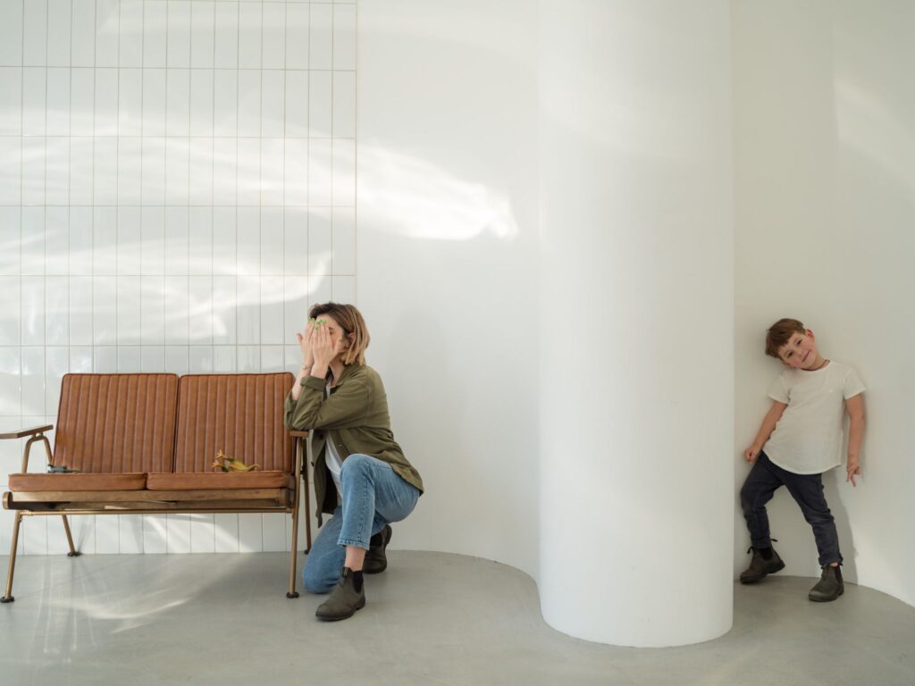 Playing hide and seek and mum sits by the sofa while a boy in a white t-shirt hides behind the wall.