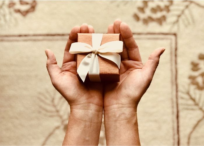 Two gift giving hands holding a small brown box tied with a white bow being given in love.