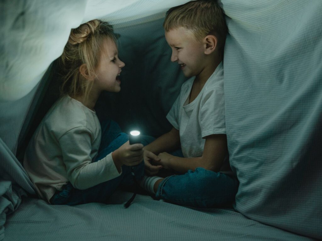 A young girl and boy having screen free time by building a fort indoors. Sitting laughing with a torch.
