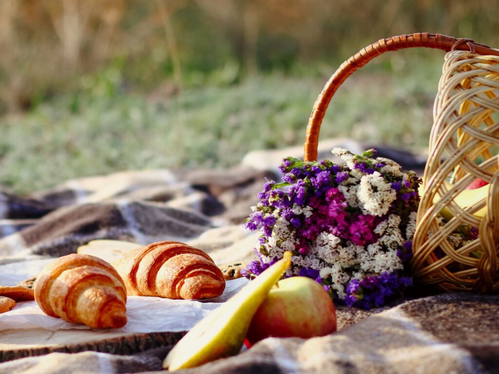 Picnic basket with food and flowers on a blanket on the grass outside.