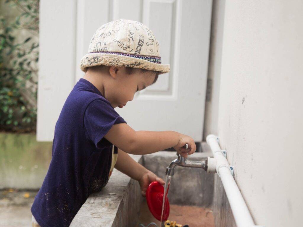 Young boy wearing a blue t-shirt and hat doing an outdoor tap activity over a sink while holding a red toy.