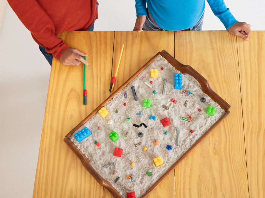 A tray with sand and plastic toys for sand tray play, sitting on a table with two kids looking.