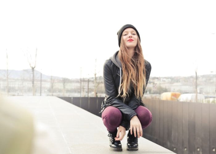 Teen girl crouched down on a structure outside with a fence behind her and trees and hills in the background.