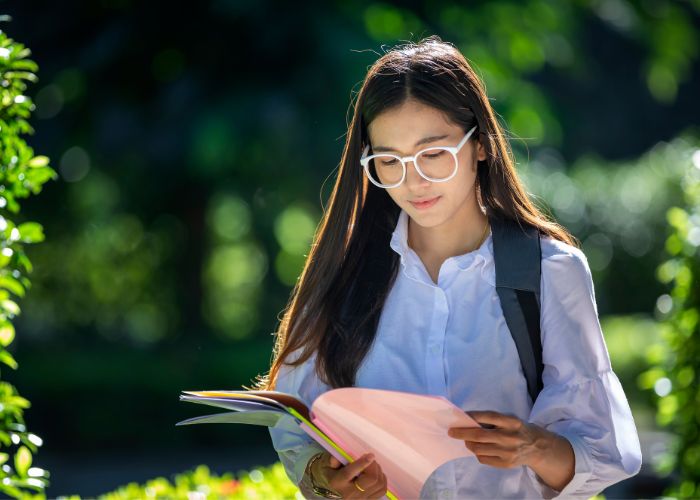 Teen girl wearing glasses and a white shirt reading a notebook as she walks through a park