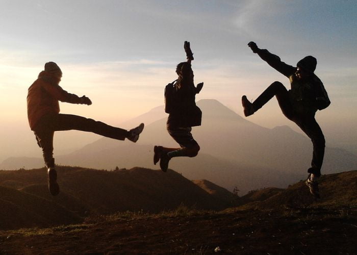 A silhouette image of three young people wearing outdoor clothing jumping and celebrating on top of a mountain range.