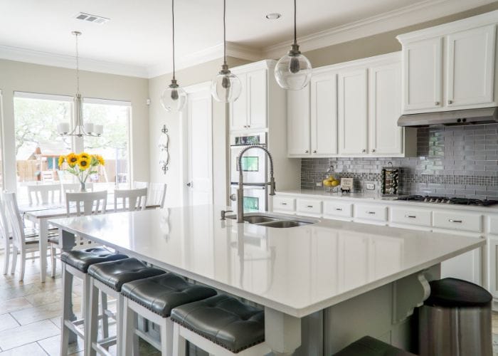 beautiful clean, white kitchen with an island bench in the middle of the room and bar stools along the side. Table and chairs in the background.
