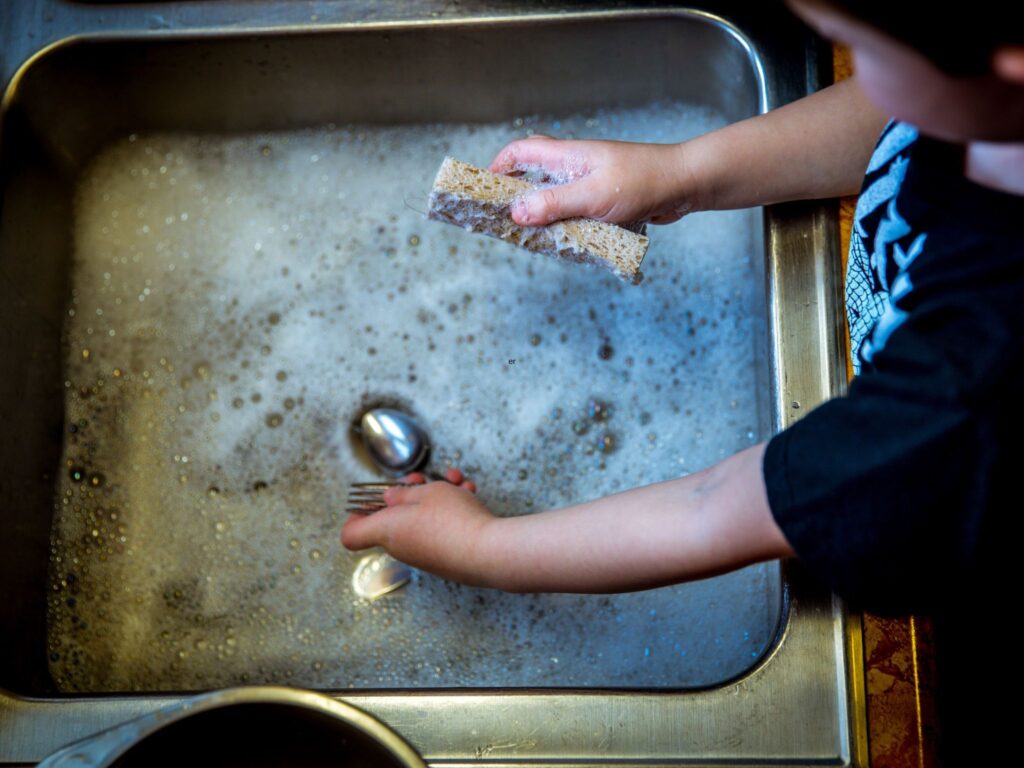 A kitchen sink with soapy water being played in by a child holding a sponge and a spoon, washing the dishes.